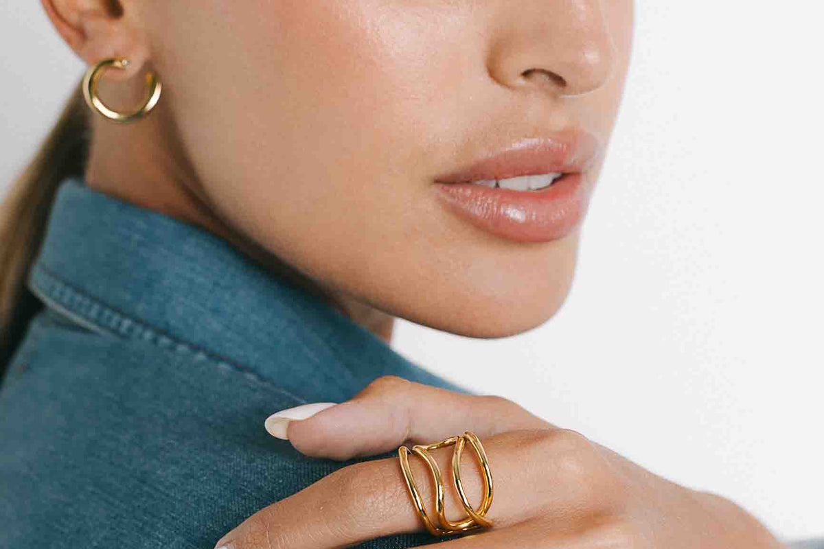 A close-up lifestyle shot of a woman wearing a chunky gold ring with three interlocking curved bands on her hand, paired with a denim shirt and a gold hoop earring against a clean white background.