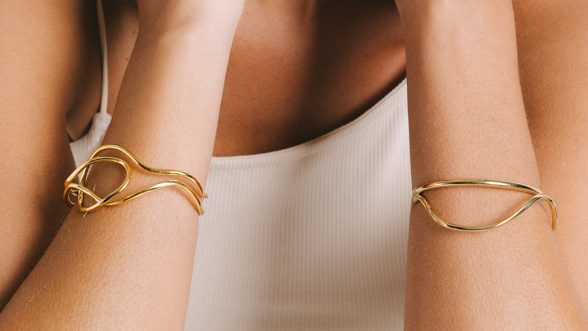 A close-up lifestyle shot of a woman wearing delicate gold jewelry, featuring a twisted wire geometric cuff bracelet on each wrist and a matching gold hoop earring.