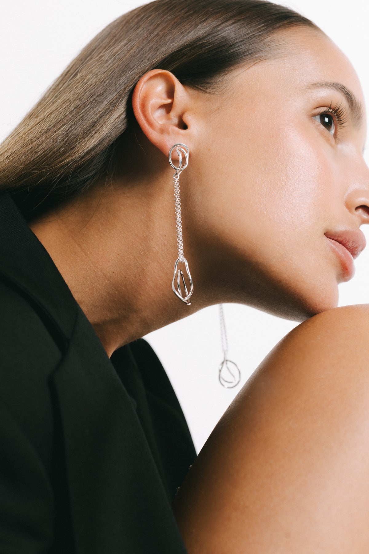 A close-up side profile of a woman wearing a long silver dangle earring with a textured chain and geometric teardrop pendant, set against a clean white background. Model wearing Fiorella Chain Drop Earrings in silver Plate by Pieretti